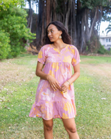 A woman wearing a pink short-sleeve tiered dress with a yellow and white floral print stands outdoors on a grassy lawn with large trees in the background.