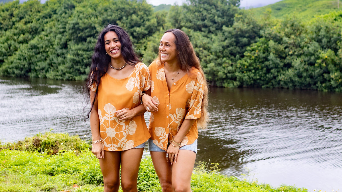 Two women smiling, wearing marigold colored tops with Hawaiian quilt design and pearl necklaces with river and trees in the background