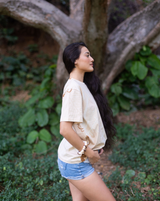 A woman with long dark hair stands outdoors in a park-like setting with a large tree trunk and green foliage behind her. She is wearing a light beige oversized t-shirt with small embroidered details and light blue denim shorts.