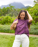 A woman wearing a purple short-sleeve t-shirt with a tonal butterfly pattern and white pants, standing outdoors in a grassy area with green foliage and mountains in the background.